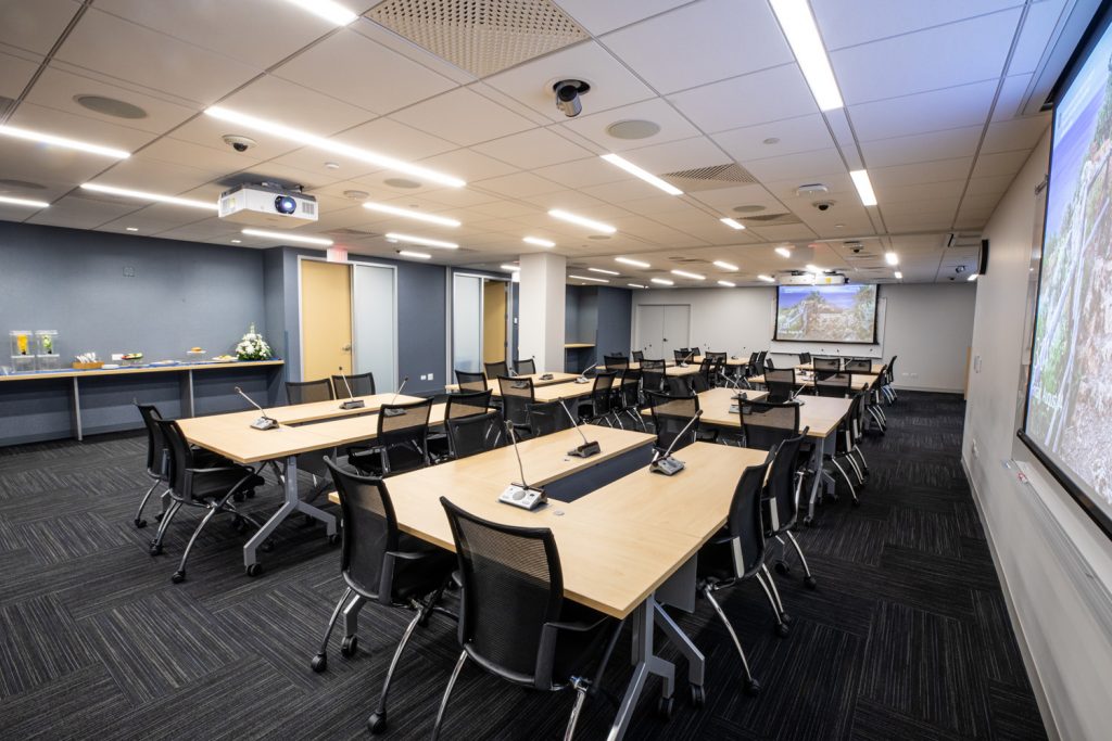 Conference Room 3AB at One Rotary Place. Photo of a large conference room with tables & chairs, microphones, and multiple projectors & screens.